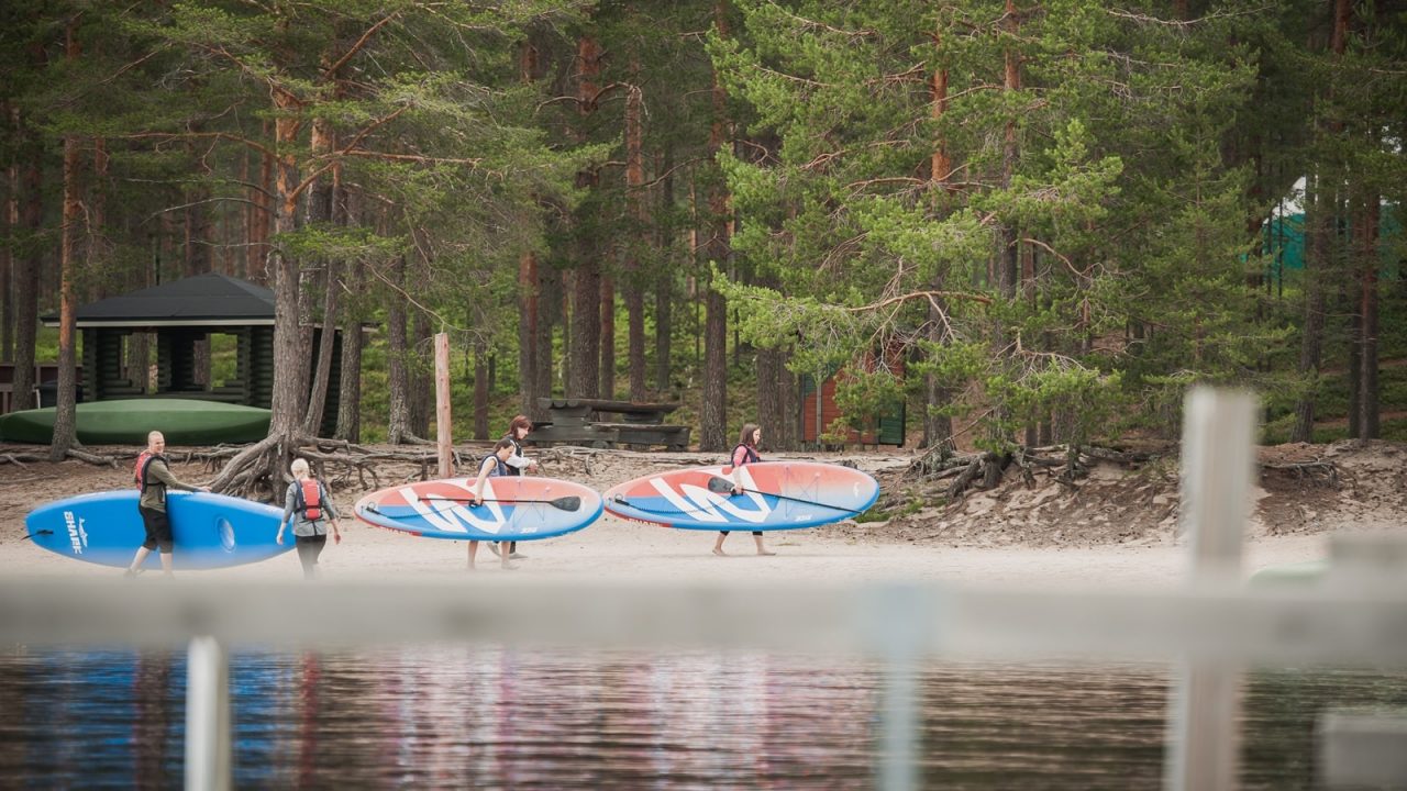 Piispalan leirintäalueen metsäinen ranta, jossa kulkee viisi suppailijaa.